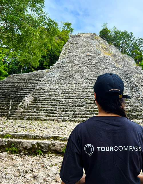 A person wearing a TourCompass T-shirt stands in front of a Mayan ruin in Mexico
