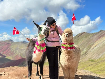 TourCompass colleague Iida stands holding two flags and an alpaca at the summit of Rainbow Mountain in Peru.
