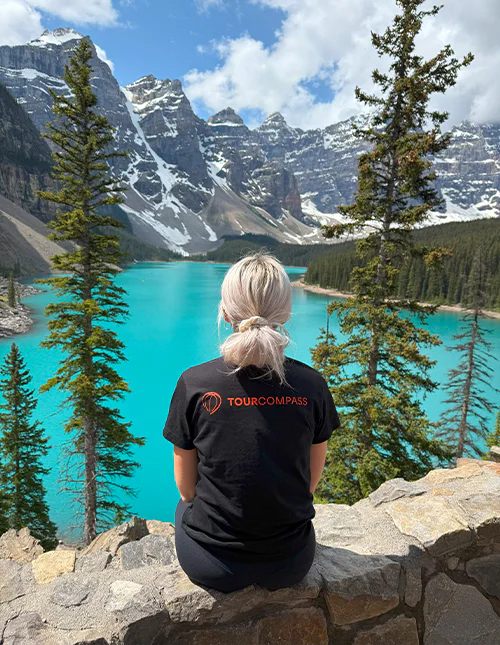 A woman in a TourCompass T-shirt looks out over a turquoise lake and snow-capped mountains in Canada