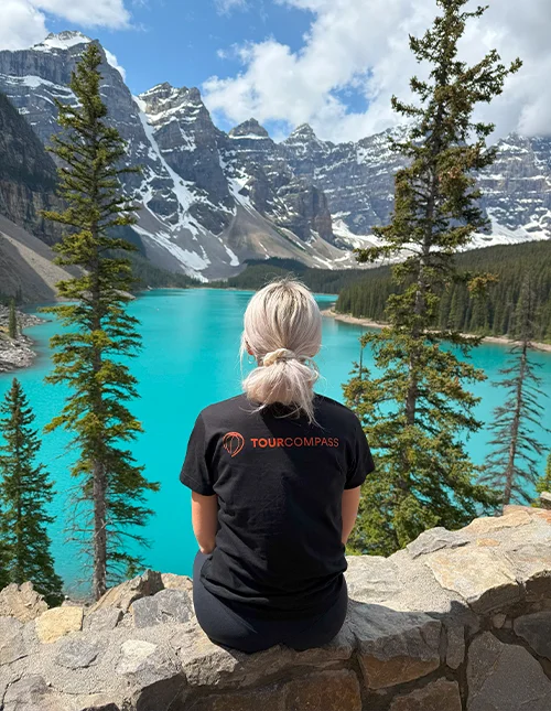 A woman in a TourCompass T-shirt looks out over a turquoise lake and snow-capped mountains in Canada
