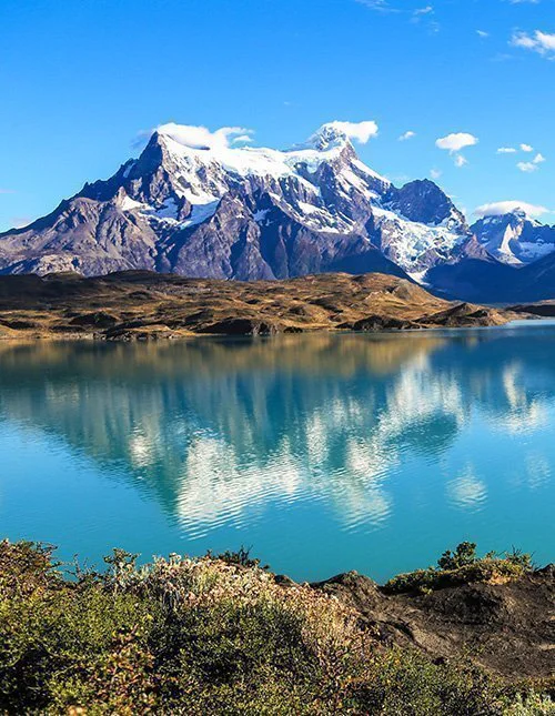 A glassy mountain lake scene, with snow-capped peaks mirrored in the turquoise water beneath a cloudless sky.