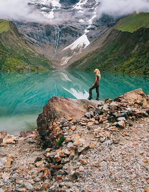 A person stands by a turquoise mountain lake, surrounded by misty, snow-capped peaks