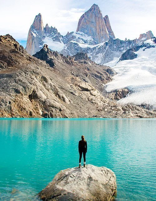 A person stands on a rocky outcrop at the edge of a turquoise glacial lake, surrounded by steep, snow-capped mountain peaks