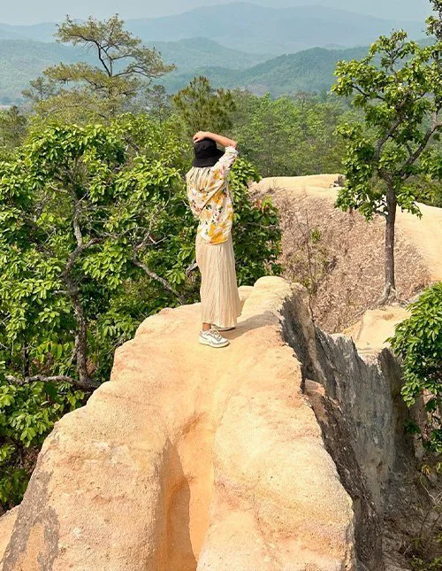 A person stands on a narrow rocky ridge overlooking green mountains